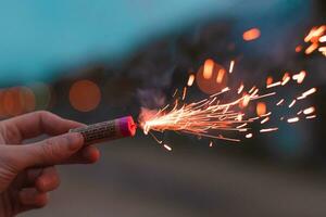 joven hombre Encendiendo arriba petardo en su mano al aire libre en noche. chico consiguiendo Listo para nuevo año divertido con fuegos artificiales o pirotécnico productos de cerca Disparo foto