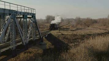 Aerial view of an old steam locomotive driving onto a railway bridge. Heritage historic steam locomotive with white smoke seen from above by drone. video