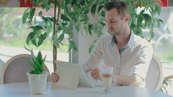 Laughing Young Customer in a Cafe Makes Video Call Using Tablet PC. Youthful Happy Handsome Man in a White Shirt Has a Coffee Break in the Daytime. Middle Angle View, Dynamic Shot