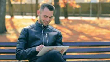 Schüler ist lesen Nachrichten oder Aufpassen Video auf Tablette Computer im das Park im Herbst. Mann im schwarz Leder Jacke ist Sitzung auf ein Bank mit groß Weiß Tablette Stk. Mitte Nahansicht Schuss