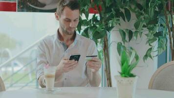joven hombre en un café hace en línea pago utilizando un teléfono inteligente y crédito tarjeta. cliente en un blanco camisa tiene un café descanso en el tiempo de día. medio de cerca Disparo video