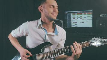 medio de cerca Disparo de un hermoso joven hombre jugando eléctrico guitarra en un oscuro ahumado habitación. activo emocional músico en un blanco camisa es grabar música a el hogar estudio en contra el negro antecedentes video