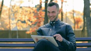 étudiant est en train de lire nouvelles ou en train de regarder vidéo sur tablette ordinateur dans le parc dans l'automne. homme dans noir cuir veste est séance sur une banc en utilisant gros blanc tablette pc. milieu fermer coup video