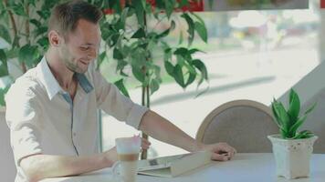 Young Happy Laughing Customer in a Cafe Makes Video Call Using Tablet PC. Youthful Handsome Man in a White Shirt Has a Coffee Break in the Daytime. Middle CloseUp View, Dynamic Shot