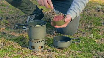 hombre en un bosque es Cocinando pasta con Enlatado estofado utilizando un pequeño cocinar colocar. turista en un caminata es preparando para el almuerzo. de cerca vista, estático Disparo video