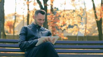 Schüler ist lesen Nachrichten oder Aufpassen Video auf Tablette Computer im das Park im Herbst. Mann im schwarz Leder Jacke ist Sitzung auf ein Bank mit groß Weiß Tablette Stk. Mitte Nahansicht Schuss