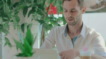 Young Happy Laughing Customer in a Cafe Makes Video Call Using Tablet PC. Youthful Handsome Man in a White Shirt Has a Coffee Break in the Daytime. Middle CloseUp View, Dynamic Shot