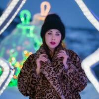 Portrait of young woman in black hat and fur coat with leopard pattern stands on background of Christmas lights decorations and looking at camera. Depth of field, selective soft focus on foreground photo