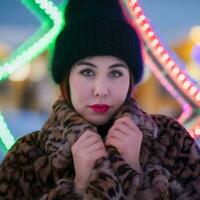 Portrait of shy young woman in black hat and fur coat with leopard pattern stands on background of Xmas lights decorations and looking at camera. Depth of field, selective soft focus on foreground photo