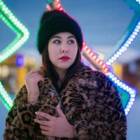 Portrait of shy young woman in black hat and fur coat with leopard pattern stands on background of Xmas lights decorations and looking away. Shallow depth of field, selective soft focus on foreground photo