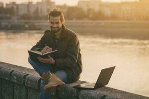 Handsome businessman using personal organizer and laptop while sitting by the river.Toned image photo