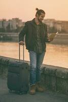 Handsome businessman with suitcase holding map of the city and standing by the river.Toned image photo