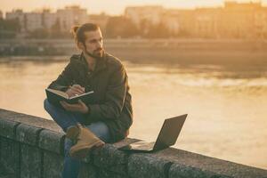 Handsome businessman using personal organizer and laptop while sitting by the river.Toned image photo