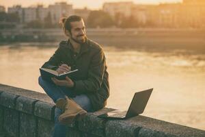 Handsome businessman using personal organizer and laptop while sitting by the river.Toned image photo