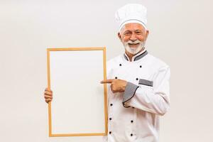Portrait of senior chef pointing at  whiteboard on gray background. photo