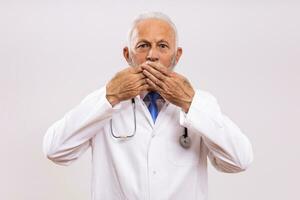 Shocked senior doctor with hands covering mouth  on gray background photo