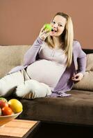 embarazada mujer sentado en sofá y comiendo manzana foto