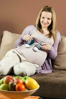 Smiling pregnant woman sitting on sofa and holding baby booties photo