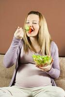 embarazada mujer sentado en el sofá y comiendo ensalada foto