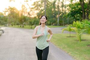 ajuste asiático joven mujer trotar en parque sonriente contento corriendo y disfrutando un sano al aire libre estilo de vida. hembra persona que practica jogging. aptitud corredor niña en público parque. sano estilo de vida y bienestar siendo concepto foto