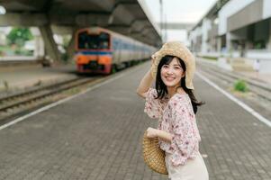 joven asiático mujer viajero con Costura cesta contento sonriente y mirando a un cámara junto a tren viniendo antecedentes. viaje viaje estilo de vida, mundo viaje explorador o Asia verano turismo concepto. foto