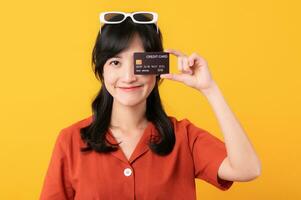 Portrait young beautiful asian woman happy smile dressed in orange clothes showing credit card isolated on yellow background. Pay and purchase shopping payment concept. photo