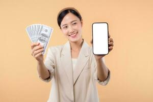 portrait of happy successful confident young asian business woman wearing white jacket holding smartphone and cash money dollar standing over beige background. millionaire business, shopping concept. photo