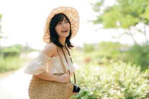 retrato de asiático joven mujer viajero con Costura sombrero y cesta y un cámara en verde público parque naturaleza antecedentes. viaje viaje estilo de vida, mundo viaje explorador o Asia verano turismo concepto. foto