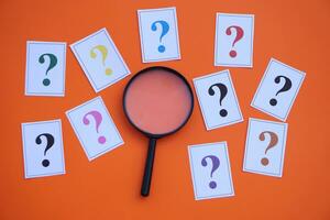 Paper cards of colorful question marks and magnifying glass on orange background. Concept. Teaching aid. Education materials for doing activity or playing investigation games about finding answers. photo