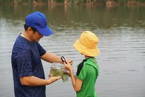 Asian father and son is exploring ecology in lake, father hold glass jar of water, son holds magnifying glass. Concept,study ecology subject explore water from nature source.Education, Life experience photo