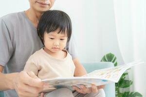 Happy Asian father relax and read book with baby time together at home. parent sit on sofa with daughter and reading a story. learn development, childcare, laughing, education, storytelling, practice. photo