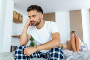 Sad thoughtful man after arguing with girlfriend.Relationship difficulties, conflict and family concept,unhappy couple having problems at bedroom. Sad guy sitting on bed,girlfriend in the background photo