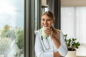 Shot of female doctor holding cup of coffee while looking forward window standing in the consultation. Smiling young woman doctor in white medical uniform and stethoscope look in distance planning. photo