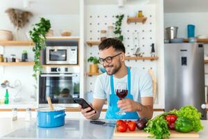 contento joven hombre preparando romántico cena buscando vegetal recetas dieta menú libro de cocina aplicación utilizando teléfono inteligente, sonriente marido participación teléfono Cocinando sano vegano comida cortar ensalada en cocina interior foto