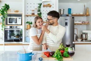 Young couple is looking at each other and feeding each other with smiles while cooking in kitchen at home. Loving joyful young couple embracing and cooking together, having fun in the kitchen photo