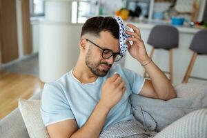Man feeling bad, sitting on the sofa with headache and applying ice bag photo