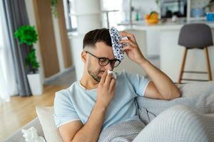 Man feeling bad, sitting on the sofa with headache and applying ice bag photo
