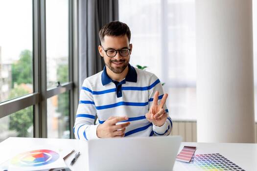 Cheerful man designer having video conference with clients, sitting at desk in front of computer, holding color palettes, gesturing and smiling, copy space photo