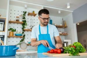 contento joven hombre el cortar Fresco vegetales o ensalada hojas, preparando vegetariano ensalada, disfrutando Cocinando solo en moderno cocina en pie a de madera encimera, pasatiempo actividad concepto. foto