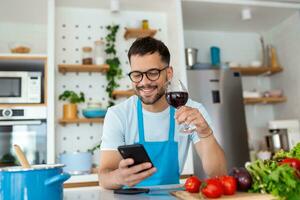 contento joven hombre preparando romántico cena buscando vegetal recetas dieta menú libro de cocina aplicación utilizando teléfono inteligente, sonriente marido participación teléfono Cocinando sano vegano comida cortar ensalada en cocina interior foto