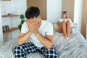 Sad thoughtful man after arguing with girlfriend.Relationship difficulties, conflict and family concept,unhappy couple having problems at bedroom. Sad guy sitting on bed,girlfriend in the background photo