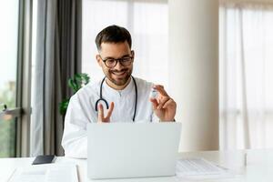 Doctor with laptop conducts an online video call and talks to patient. Male doctor working at office desk, office interior on background photo