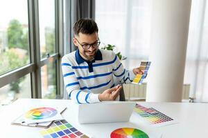 web designer working from home, sitting at table with modern laptop, looking at color samples, happy man freelancer enjoying his remote job, copy space photo