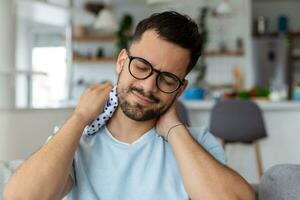 Neck Pain. Young Man Massaging Aching Neck Suffering From Ache Sitting On Couch In Living Room At Home. Osteoarthritis, Health Problem Concept. photo