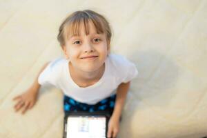 retrato de chica pequeño niña jugando en digital tableta mientras sentado a hogar en vivo habitación. foto