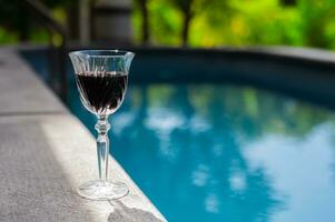 A glass of red wine puts beside outdoor swimming pool to enjoy and relaxing moment. photo