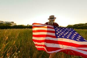 hermosa joven niña participación un americano bandera en el viento en un campo de centeno. verano paisaje en contra el azul cielo. horizontal orientación. foto