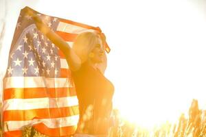 hermosa joven niña participación un americano bandera en el viento en un campo de centeno. verano paisaje en contra el azul cielo. horizontal orientación. foto