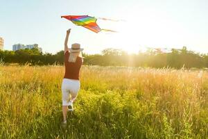 Beauty girl running with kite on the field. Beautiful young woman with flying colorful kite over clear blue sky. Free, freedom concept. Emotions, healthy lifestyle photo