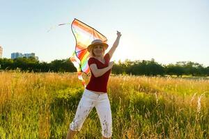 Beauty girl running with kite on the field. Beautiful young woman with flying colorful kite over clear blue sky. Free, freedom concept. Emotions, healthy lifestyle photo